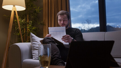 A man sitting on a cozy sofa in living room, attentively reviewing documents. Working on bills, financial planning, or reports.