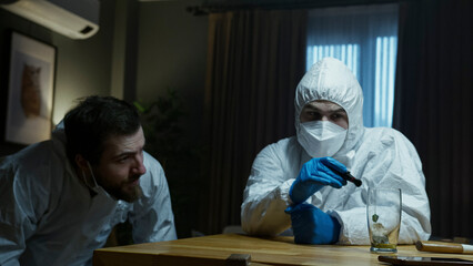Forensic investigator in a white hazmat suit and blue gloves closely examines evidence at a dimly lit crime scene.
