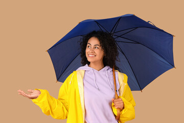 Happy young African-American woman in raincoat with umbrella on beige background