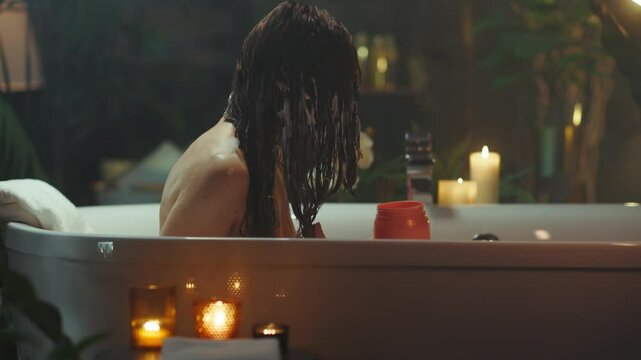 A woman with wet, dark hair relaxes in a white bathtub, applying hair product amidst the warm glow of candles and lush plants. The intimate scene evokes deep self-care and a serene beauty routine.