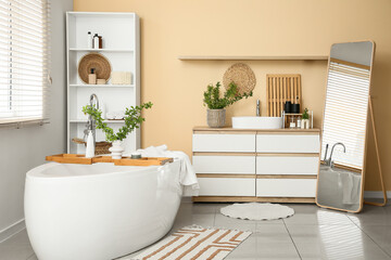 Interior of stylish bathroom with bathtub, shelving unit, mirror and green blossoming branches in...