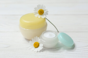 Jars of chamomile cosmetic cream and flowers on white wooden background