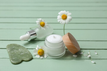 Different chamomile cosmetic products with guasha tool and flowers on green wooden background