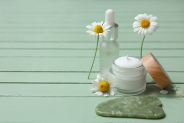 Different chamomile cosmetic products with guasha tool and flowers on green wooden background