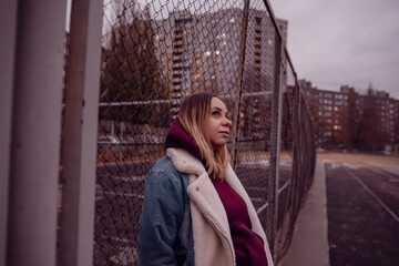 A young woman in a cozy jacket leans against a chain link fence at an urban park during autumn in...