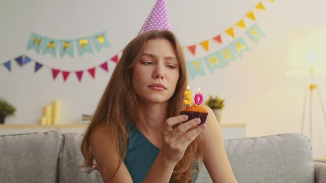 A young sad woman with red hair wears a party hat while holding a cupcake with candles at her birthday celebration indoors. She reflects on her milestone birthday surrounded by decorations.