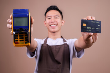Happy Young Asian Man Wearing Brown Apron Showing Credit Card And EDC Machine Over Beige Studio Wall
