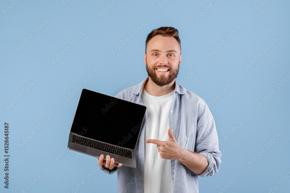 Poster positive young man pointing at empty laptop screen with space for your website design over blue stud