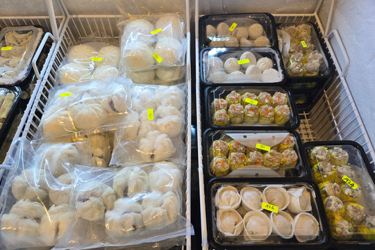 Assorted frozen dim sum including bao buns, dumplings, and siu mai packed in transparent plastic and black trays displayed neatly in a supermarket freezer.