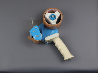 Parcel tape dispenser with brown roll of tape on grey background, Handheld dispenser with roll of adhesive tape isolated on grey. Device for preparing goods for shipment. 