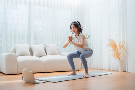 Young Asian beautiful woman doing aerobic exercise at home. Attractive girl doing  workout by squatting, follow instructions video from online trainer for health in house