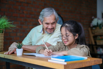 Young asian schoolgirl is doing her class homework assignment at home with advice from grandfather for education and study