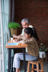 Young asian schoolgirl is doing her class homework assignment at home with advice from grandfather for education and study