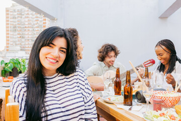 Young latin woman smiles at camera, on terrace at lunch with friends where they sing and play guitar