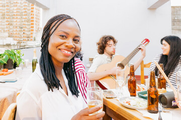 Young black woman smiles at camera holding a glass cup, on rooftop, as her friends play guitar