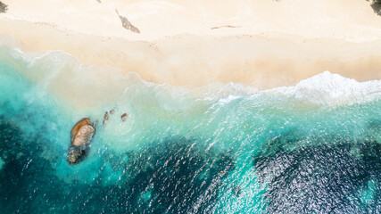 Aerial view of gentle turquoise waves brushing the sandy shore with a glimpse of rocks beneath the water. Praslin, Seychelles.