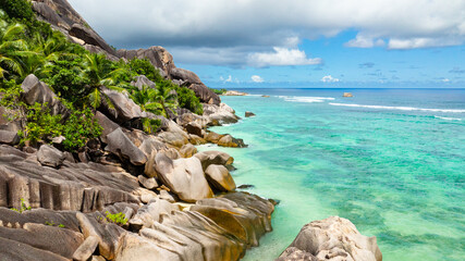 Granite rocks resting on a tropical coastline with turquoise waters and lush green vegetation. La Digue, Seychelles.