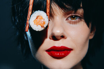 Young woman with black hair holding sushi near her face, showcasing fresh salmon, bold red lips, and dark background for a vibrant food concept.