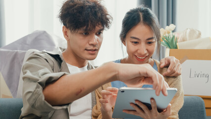 Young happy Asia couple sit on sofa couch use digital tablet online shopping furniture from decoration store in living room at home. Husband wife move new house, Apartment Rent And Ownership Concept.