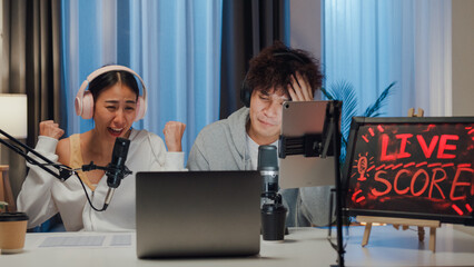 Young happy Asian couple wear headphone sit on desk online live streaming football live score looking at camera watch soccer sport game on laptop talk with follower in small studio at home at night.