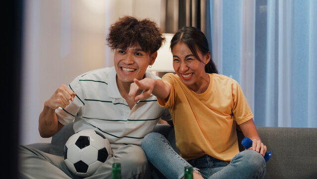 Asian new marriage couple sit on sofa excited cheering watching football sport game on television in living room at home. Young couple sport fans hold football watching online soccer on TV.