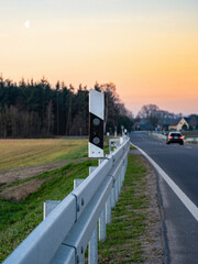 Blick auf die Leitplanke und einen Leitpfosten einer Landstraße am Morgen, ein kleines Haus und ein Auto unscharf im Hintergrund, vertikal 