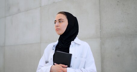 Muslim woman in hijab, white jacket holding black book, turning head sideways against concrete wall background