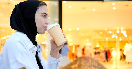 Young muslim woman wearing a hijab enjoys a coffee while using her smartphone in a brightly lit shopping mall, showcasing a blend of modern life and tradition. Free Islamic woman in a shopping center