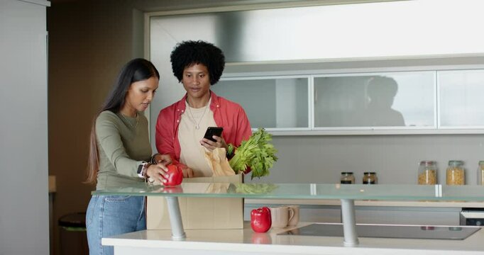 Diverse couple unpacking red pepper and olive oil from bag on kitchen island while checking recipe