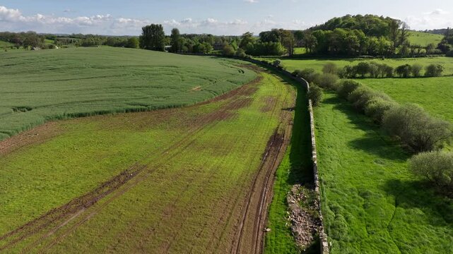 Caledon Border Wall, County Tyrone, Northern Ireland, May 2023. Drone aerial pushing towards and ascending over historic divide wall, vibrant farmland and lush greenery.