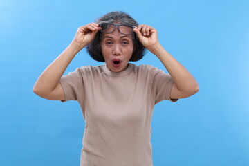 Naklejka premium Gray-haired Asian old woman, wearing glasses, open mouth, looks surprised and amazed looking at something in front while holding glasses, standing isolated on blue background.
