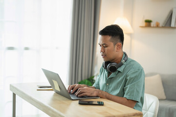 Obraz premium Young man working on laptop at home office with headphones in cozy environment, engaged in online activities while sitting at wooden desk