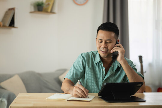 Professional man engaged in phone conversation while taking notes at home office desk, showcasing modern workspace and productivity concept in casual attire