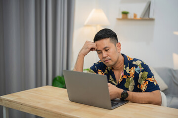 Focused man working from home with laptop in stylish casual shirt while deep in thought in modern interior setting with ambient lighting