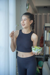 Asian woman in sportswear standing eating salad for diet in living room