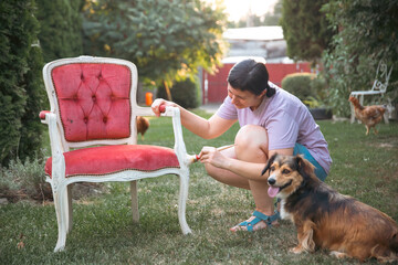 Woman painting a wooden furniture while sitting with her dog in the garden. DIY concept.