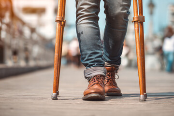 A person walking with crutches wearing jeans and brown leather boots on a wooden walkway outside city