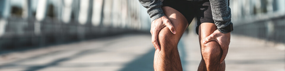 Man holding his knees in pain on a bridge after exercising or running outdoors in the sunlight