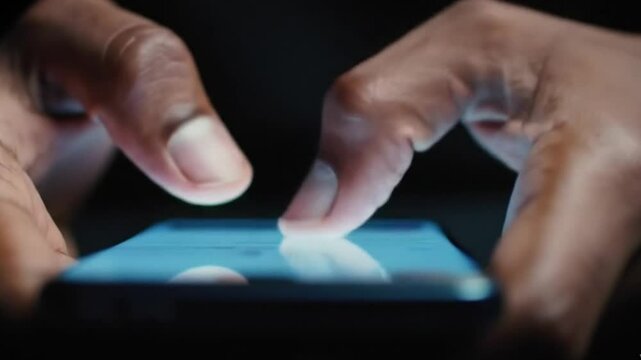 close-up shot of a man's fingers typing quickly on a smartphone screen in a dark environment. The only light source is the glow from the phone screen, casting soft highlights on his fingertips