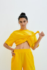 young woman in yellow casual outfit lifting shirt indoors, minimalistic background, natural look, stylish sporty clothing, relaxed modern vibe