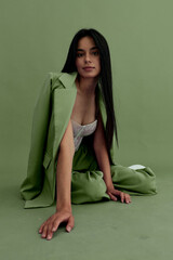 Young woman with long black hair wearing a green suit and light lace top poses confidently against a matching green studio backdrop, showcasing modern fashion style.