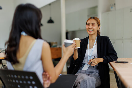 Two happy asian women holding coffee cup work in the office, talking or casual meeting during coffee break
