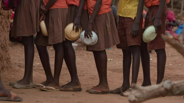African children stand in line with bowls in their hands to get portion of food during break between classes. Slow motion. Close-up of schoolchildren's feet in flip-flops