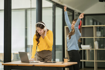 Asian woman wearing headphones standing and dancing celebrating, showing her appreciation, joy, office room