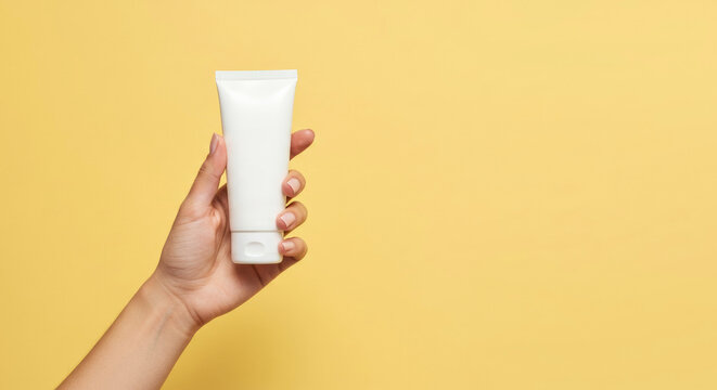 Hand holding a white tube of cream against a bright yellow background in a indoor setting