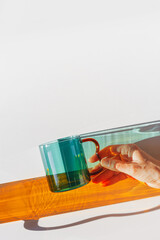 Vibrant still-life with colorful transparent glass mug or cup in shades of green teal and orange. Sunlight dynamic shadows and reflections on white, minimal aesthetic design, table setting