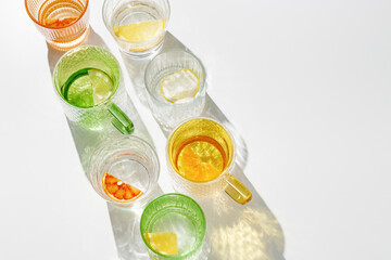 Vivid Glasses with cold detox water as summer still life pattern, shadow and sunlight reflection, closeup glass cups of infused fruit water, slises of lemon, orange, tangerine, healthy summer drink