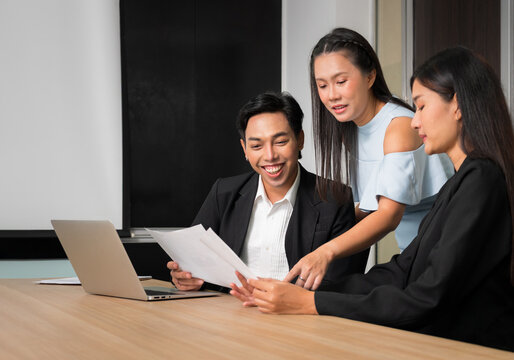 Three young professional collaborating around laptop and documents in modern office while reviewing business materials together at conference table room. Teamwork and business collaboration concept - Powered by Adobe