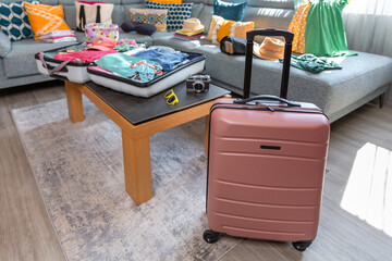 Luggage and clothing scattered across the sofa in a living room as part of travel preparation. Open suitcase over table with colorful summer clothes, ready for vacation. Copy space available on left.