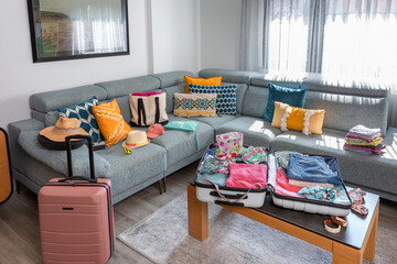 Open suitcase with summer clothes and accessories placed on coffee table and sofa in living room, symbolizing upcoming vacation and travel preparations